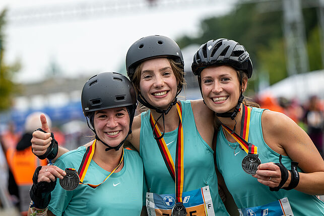Three young women are laughing into the camera. They have medals hanging around their necks and they all have the same shirt and a black helmet.