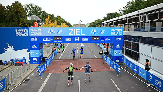 Two inline skaters cross the finish line holding hands. Behind them, other skaters cross the finish line.