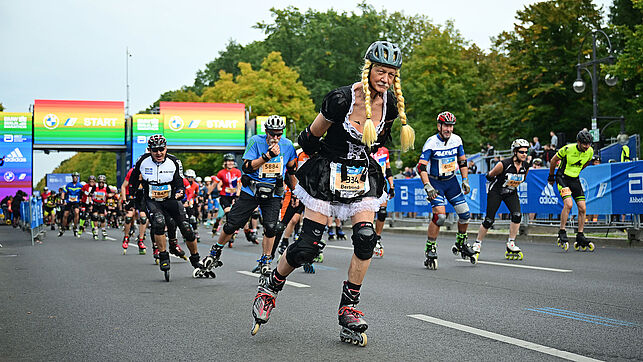 A male skater wearing a wig with long blond braids has just passed through the start gate in rainbow colours. His disguise also includes a short dress.
