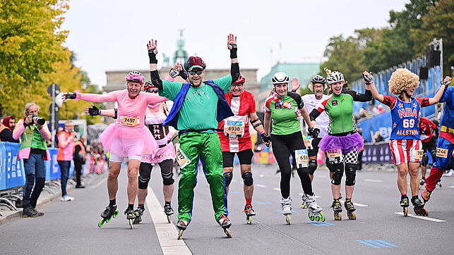 A jubilant group of skaters dressed in very different outfits ride side by side on the finishing straight. One male skater is wearing a pink ballet dress.